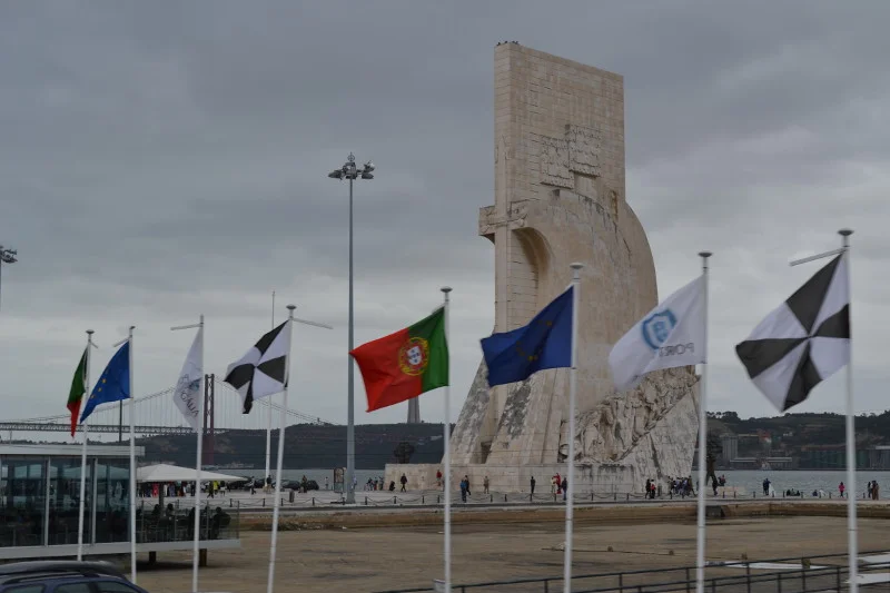 Monumento aos Descobrimentos, às margens do rio Tejo, em Lisboa (ao fundo, ponte 25 de abril). O monumento tem a forma de uma caravela estilizada, com o escudo de Portugal nos lados e a espada da Casa Real de Avis sobre a entrada. D. Henrique, o Navegador, ergue-se à proa, com uma caravela nas mãos. Em duas filas descendentes, de cada lado do monumento, estão as estátuas de heróis portugueses ligados aos Descobrimentos. Na face ocidental encontram-se o poeta Camões, com um exemplar de Os Lusíadas, o pintor Nuno Gonçalves com uma paleta, bem como famosos navegadores, cartógrafos e reis.