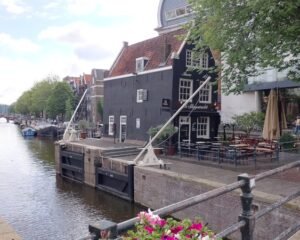 De Sluyswacht (bar em antiga torre do canal que entortou bastante com os anos) e canal Oudeschans com Montelbaanstoren ao fundo (torre de defesa do século 16, outrora parte das muralhas da cidade, com sinos e um relógio adicionado em 1606). Foto: ViniRoger